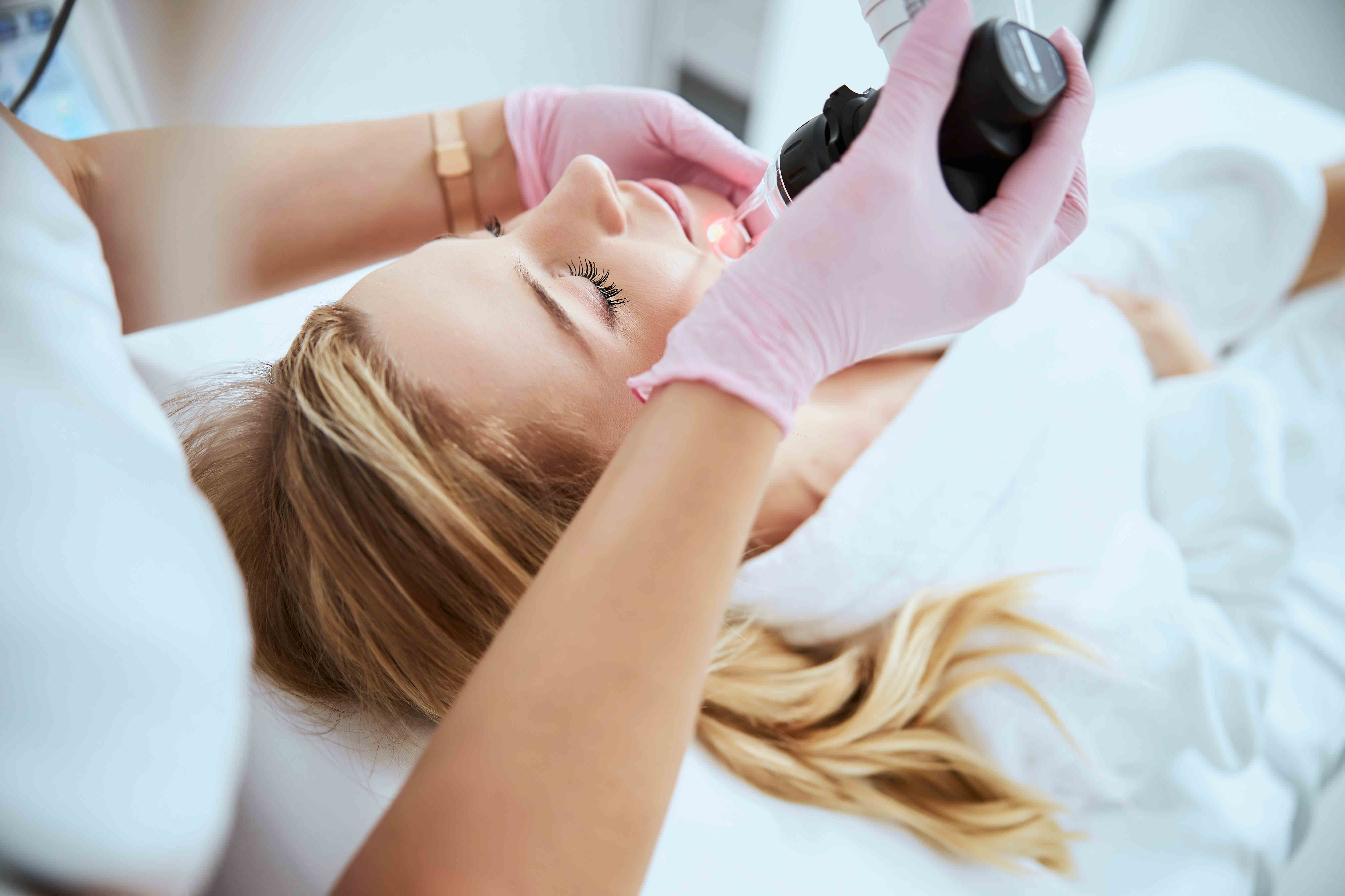 Woman receiving a facial treatment with a handheld device by a professional wearing pink gloves.