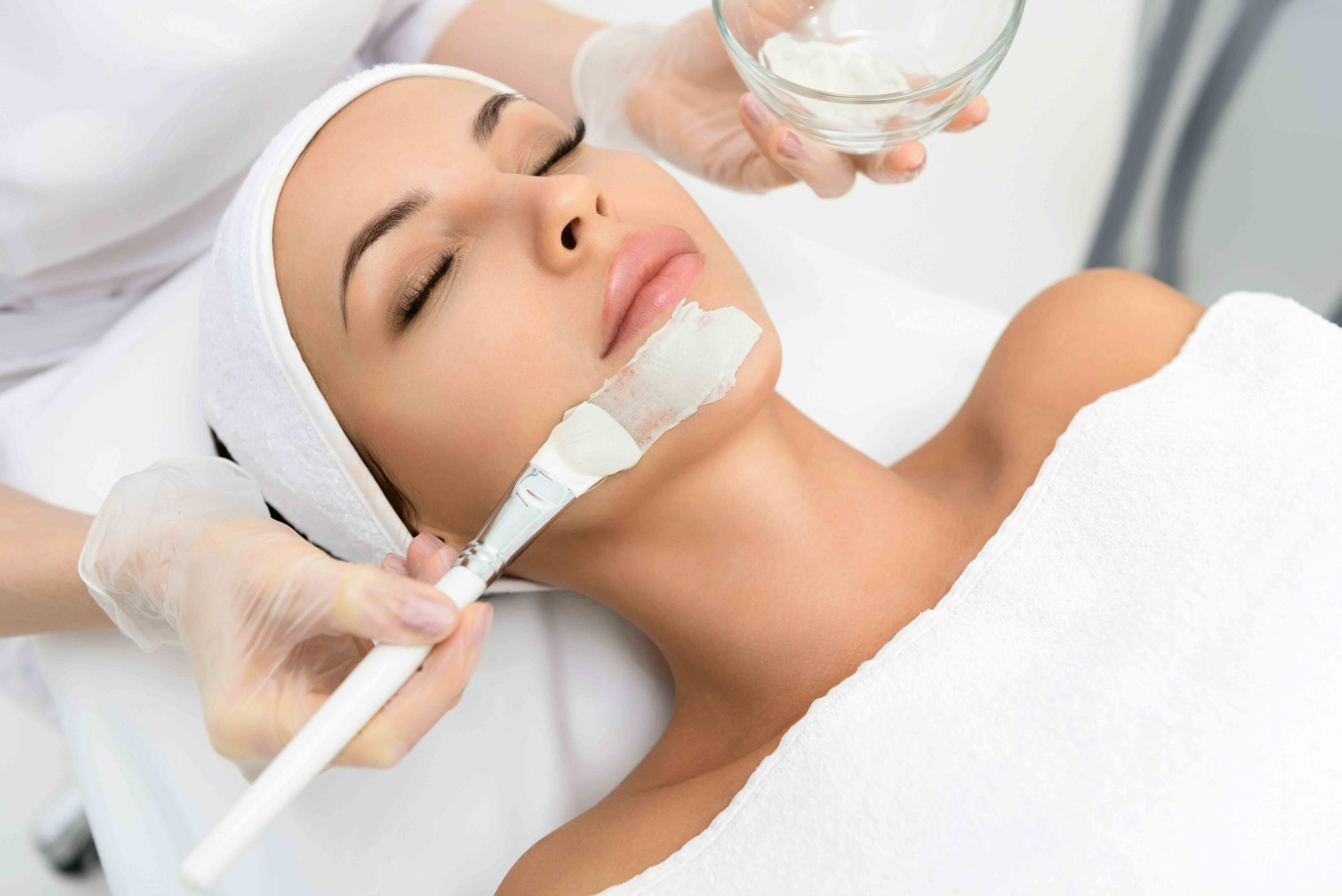 A woman with eyes closed receiving a white facial mask applied on her chin at a spa.