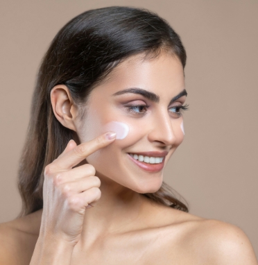 Smiling woman applying cream to her cheek with her finger against a plain beige background.